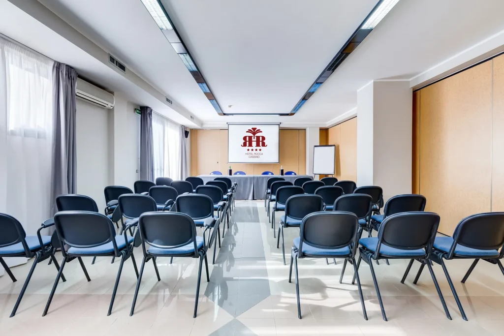 Image of a meeting room with theater-style seating. The chairs are blue, and the room is illuminated by natural light coming through a large window.