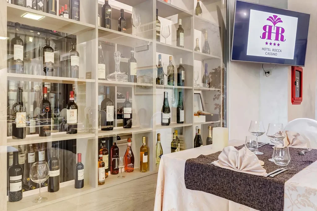 Dettaglio del ristorante: a sinistra è visibile una vetrina che espone diverse bottiglie di vino, a destra un primo piano di un tavolo apparecchiato con tovaglia beige e marrone, bicchieri da vino e da acqua e posate