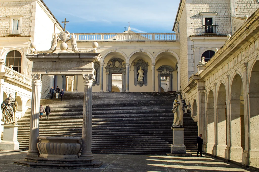 Image of the entrance to Montecassino Abbey: a central staircase, side porticos, and several sculptures decorating the area are visible.