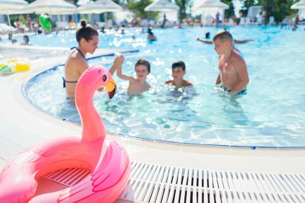Image of a flamingo-shaped swim ring in the foreground, and in the background a woman playing in a pool with three children.