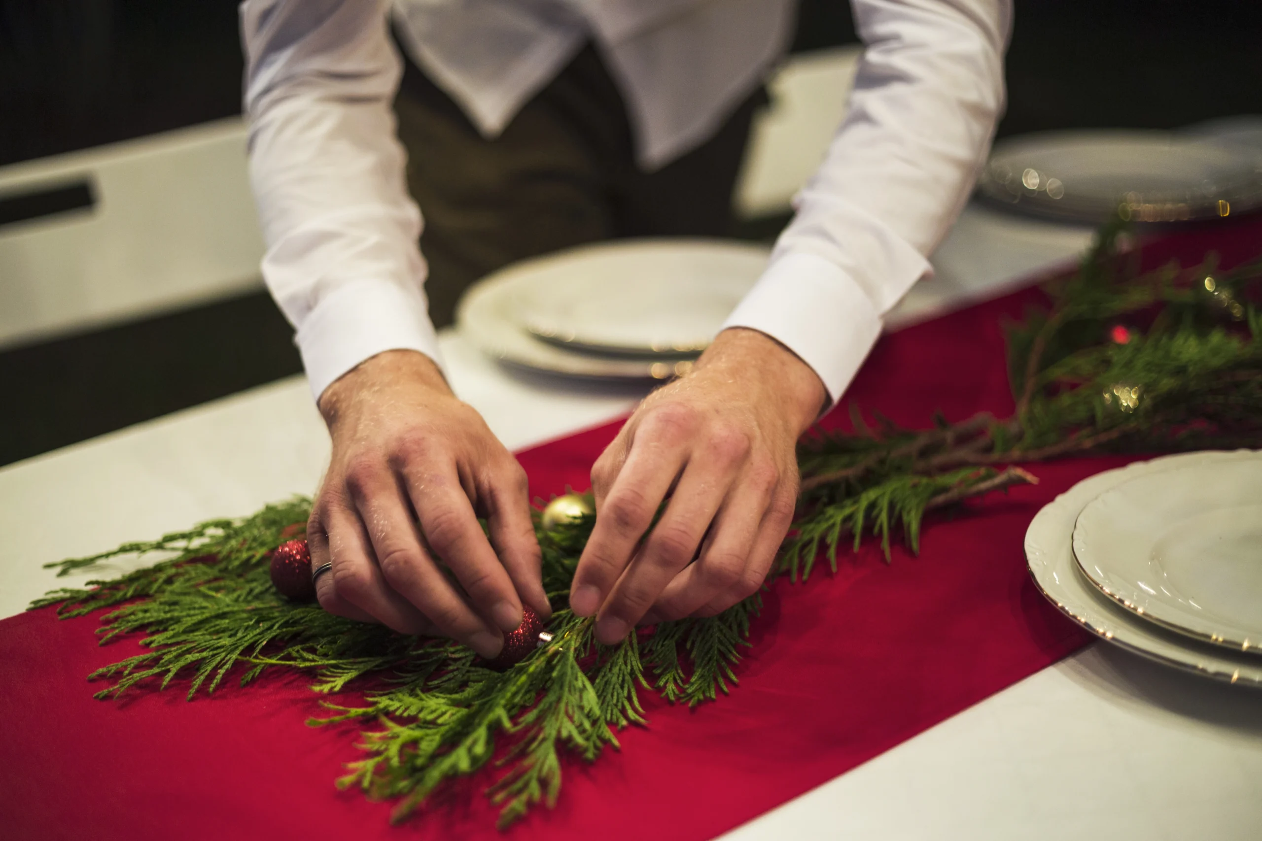 nell'immagine, due mani allestiscono una tavola per le festività natalizie: un runner rosso è posato sulla tavola e, sopra, una decorazione con rami di abete